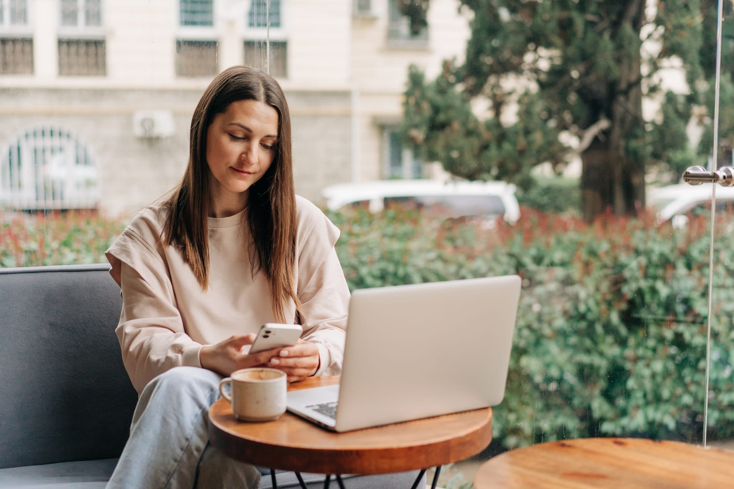 A woman at a café table typing on her phone, a laptop and a coffee in front of her