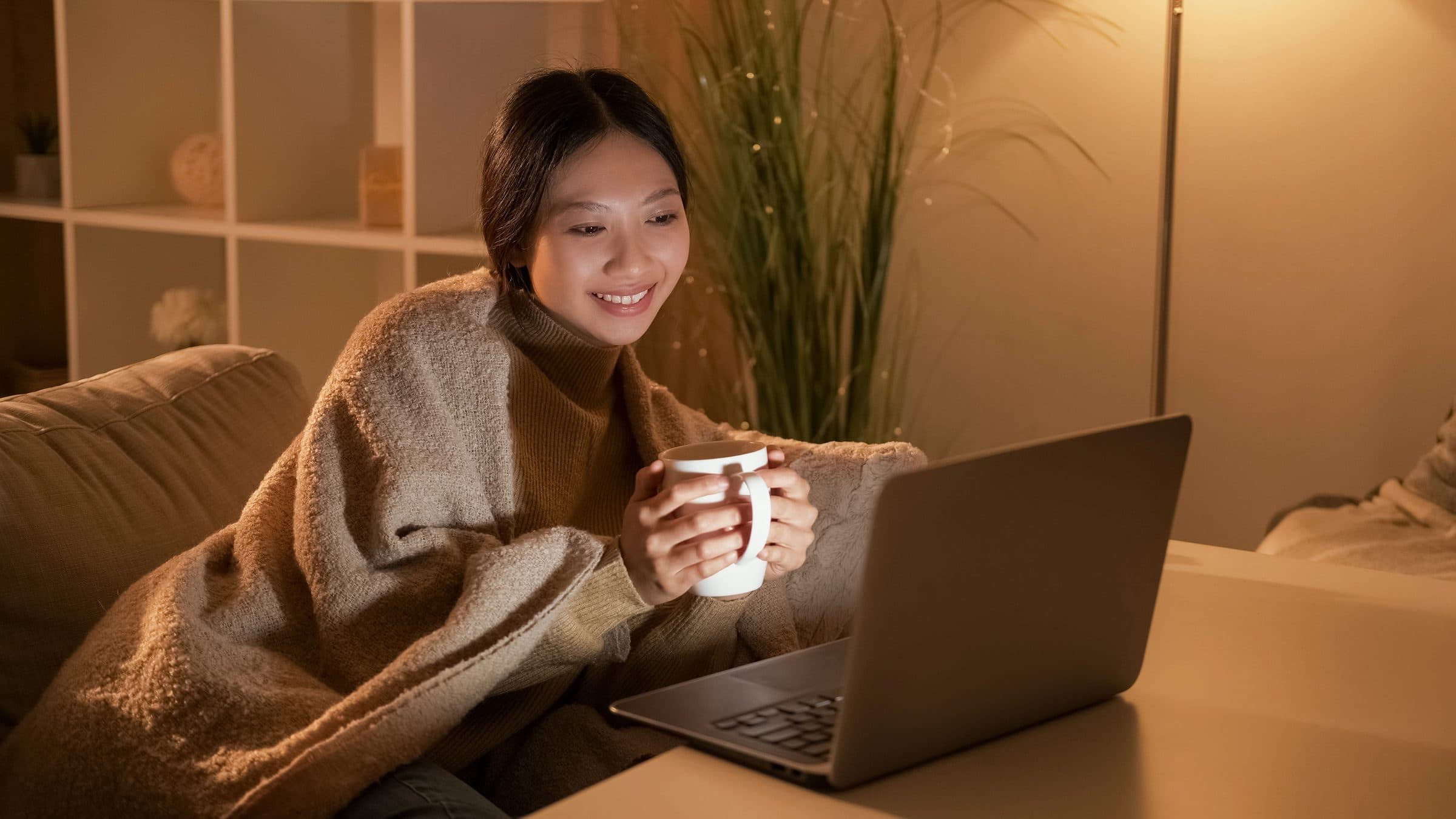 A woman wrapped in a blanket with a mug of tea, smiling at her laptop on an evening at home