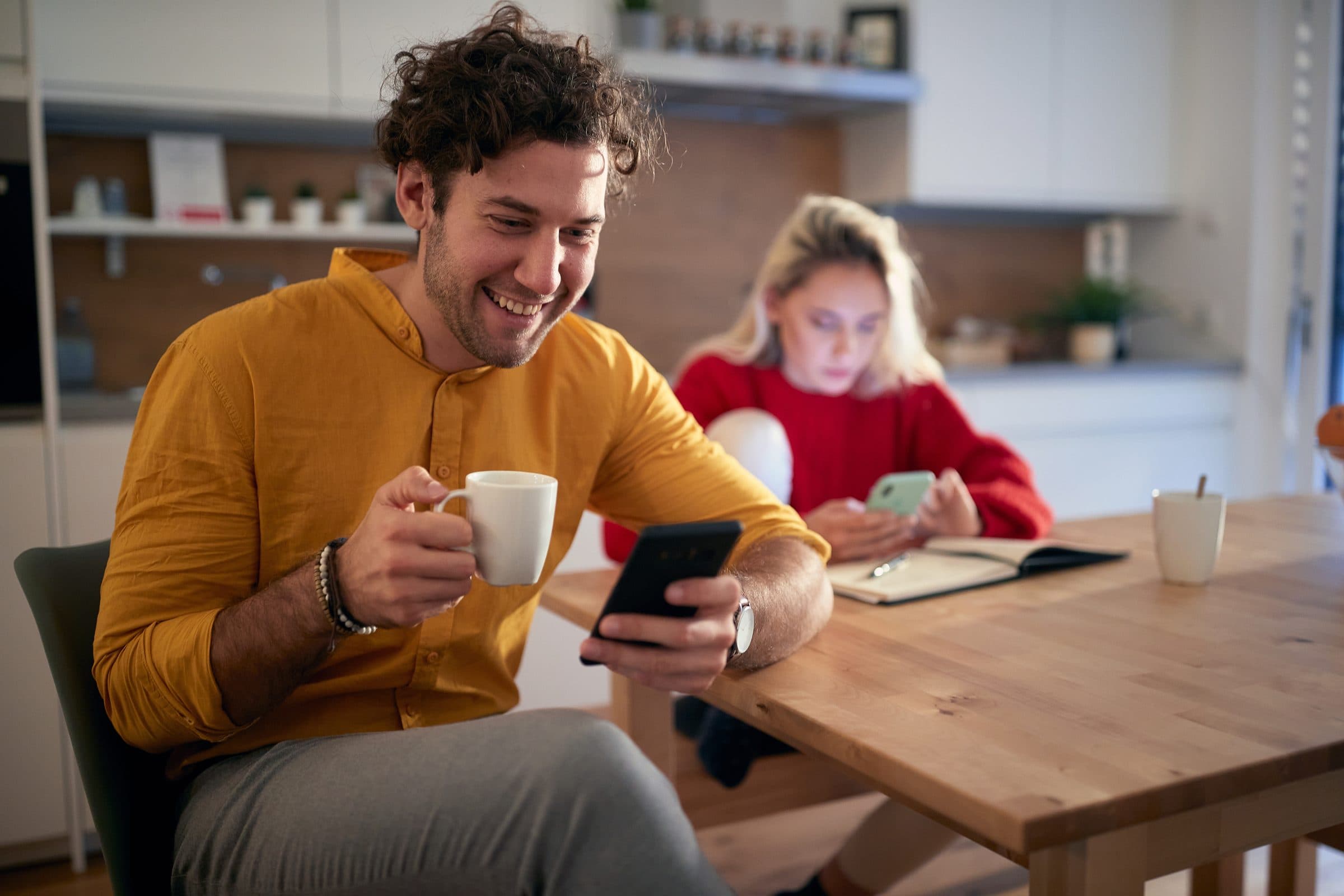 A man with a coffee smiling at his phone at a kitchen table
