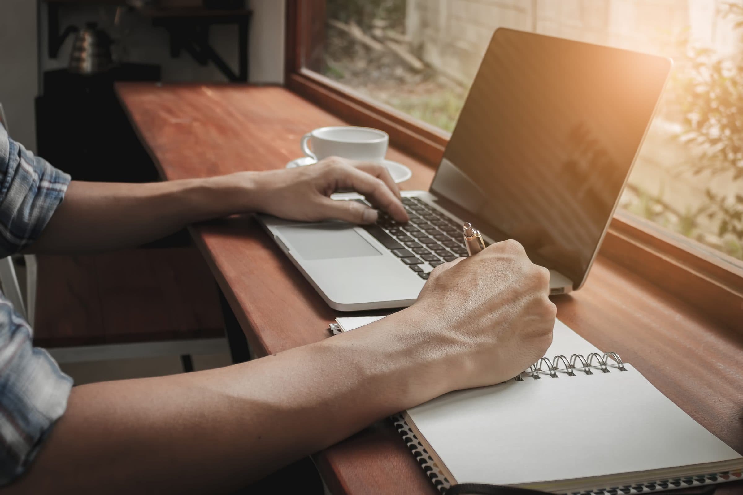 Hands typing on a laptop and writing in a notebook at a wooden desk by a window